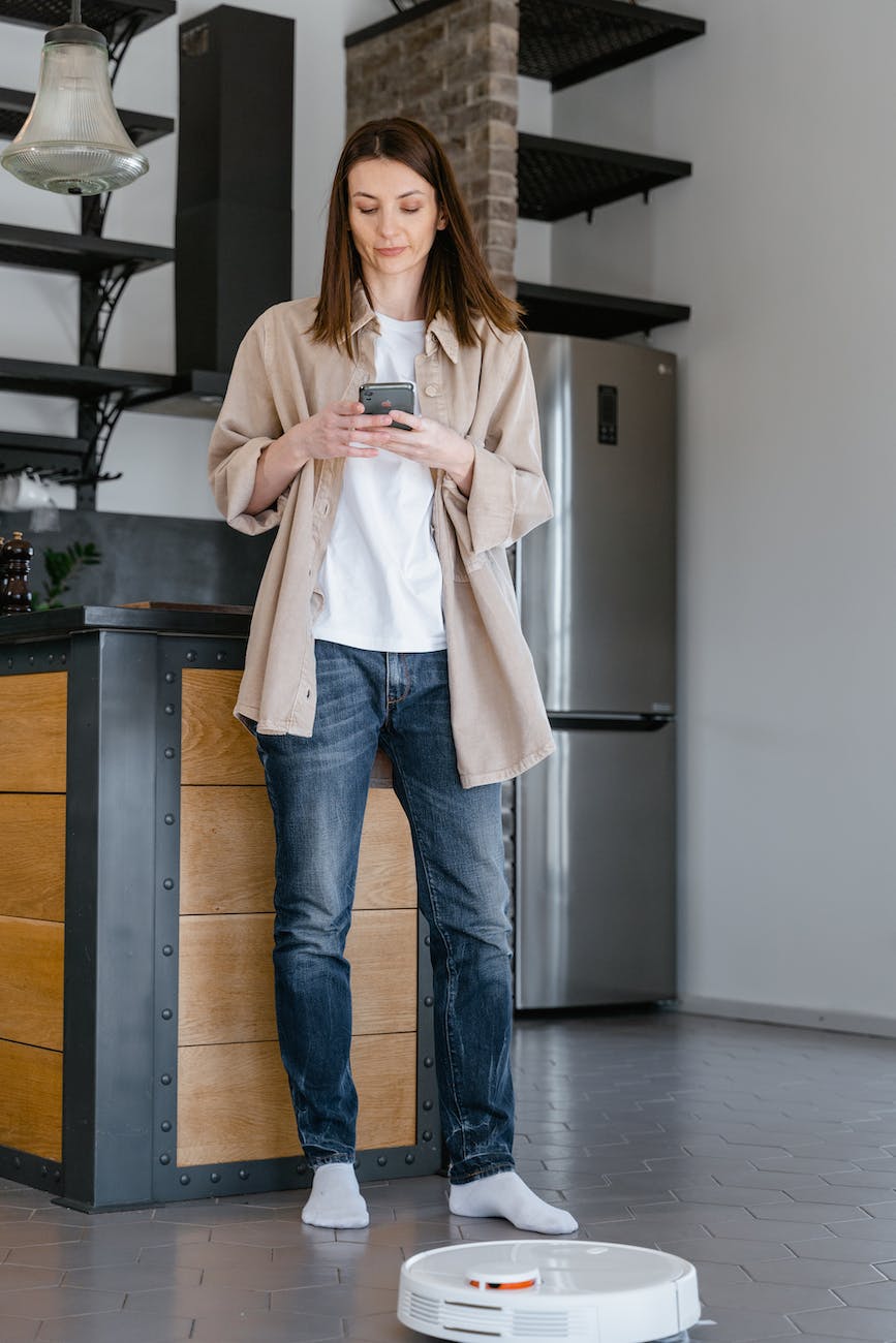 woman in beige long sleeve shirt and blue denim jeans standing beside kitchen counter while texting